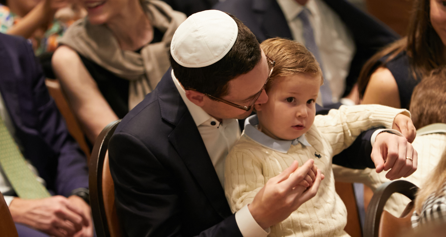 An adult wearing a kippah warmly embraces a young boy amid a gathering, both dressed in formal attire.
