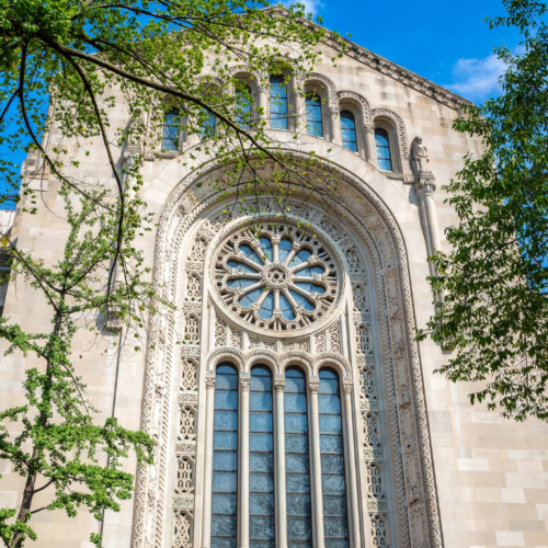 Large arched stained-glass window on the historic façade of Temple Emanu-El, framed by green tree branches with a backdrop of blue sky.