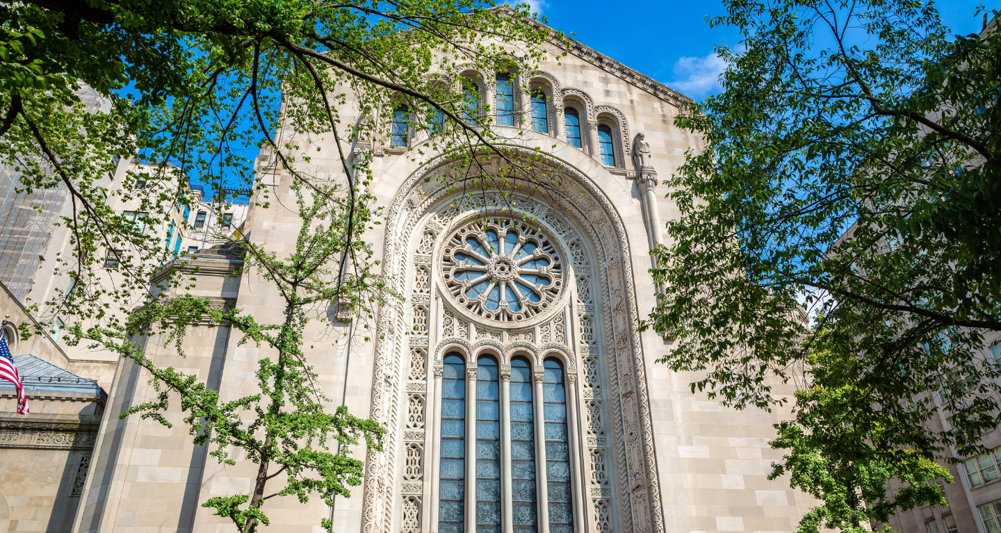 Large arched stained-glass window on the historic façade of Temple Emanu-El, framed by green tree branches with a backdrop of blue sky.