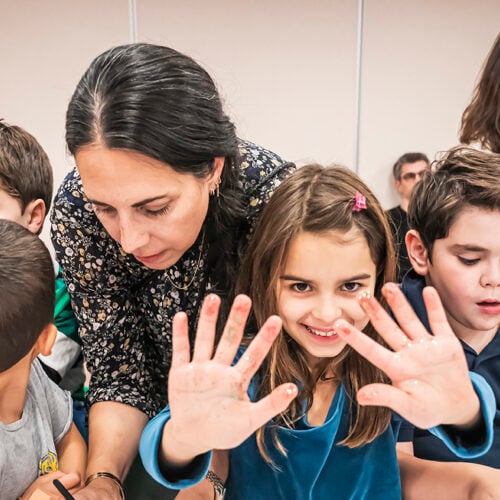 Smiling girl displaying her painted hands in a bright classroom at Temple Emanu-El, with children and adults gathered around her.