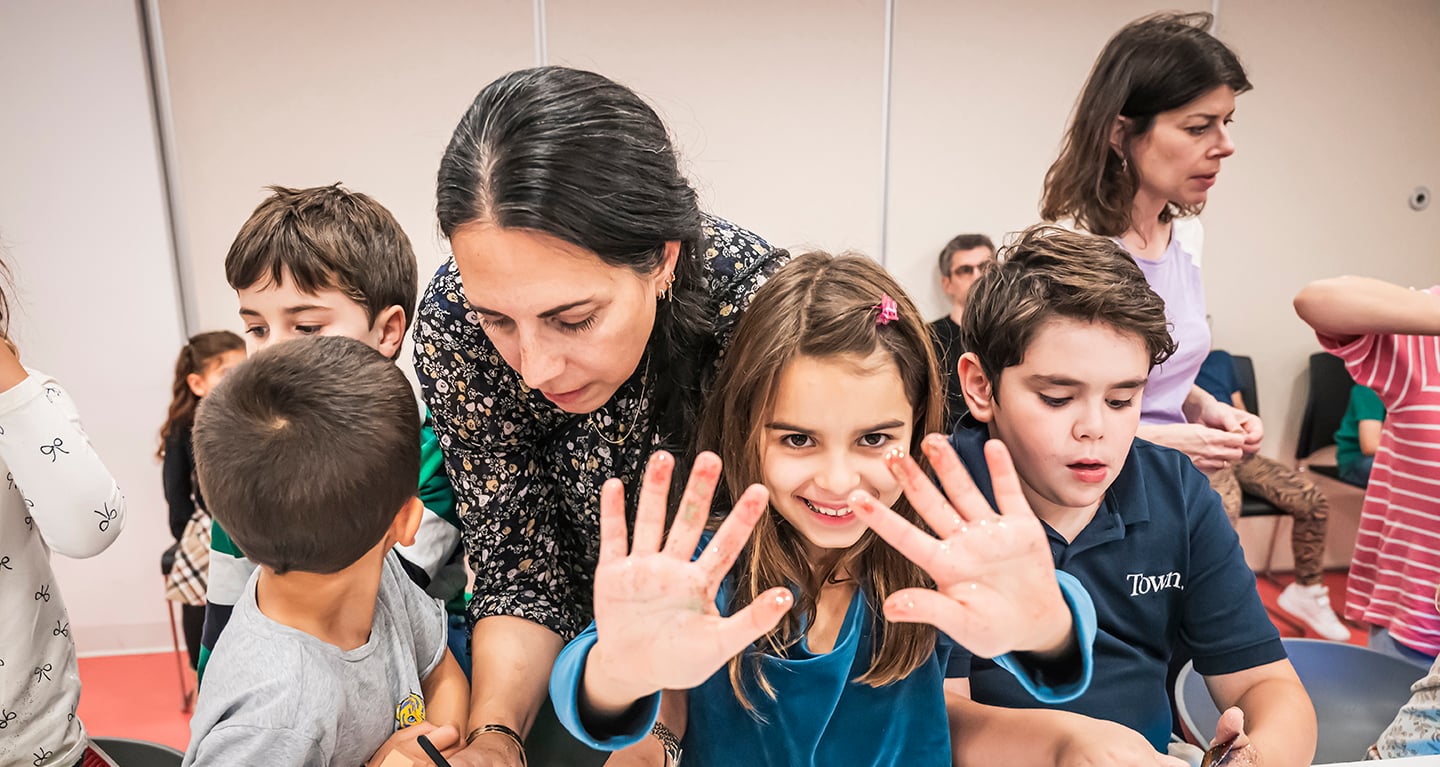 Smiling girl displaying her painted hands in a bright classroom at Temple Emanu-El, with children and adults gathered around her.