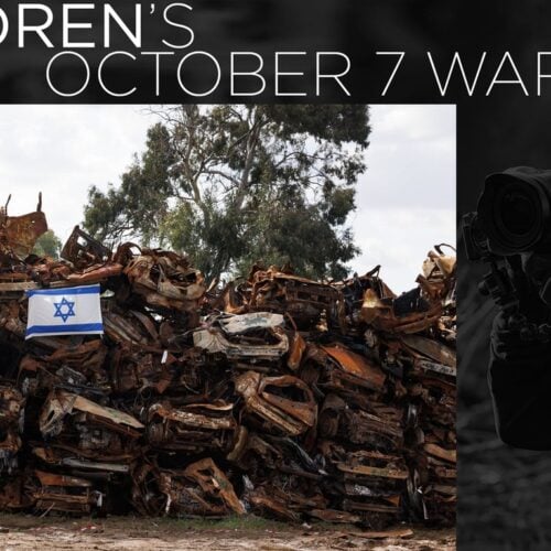 A group of damaged vehicles with an Israeli flag nearby, as a photographer documents the scene on the right side.