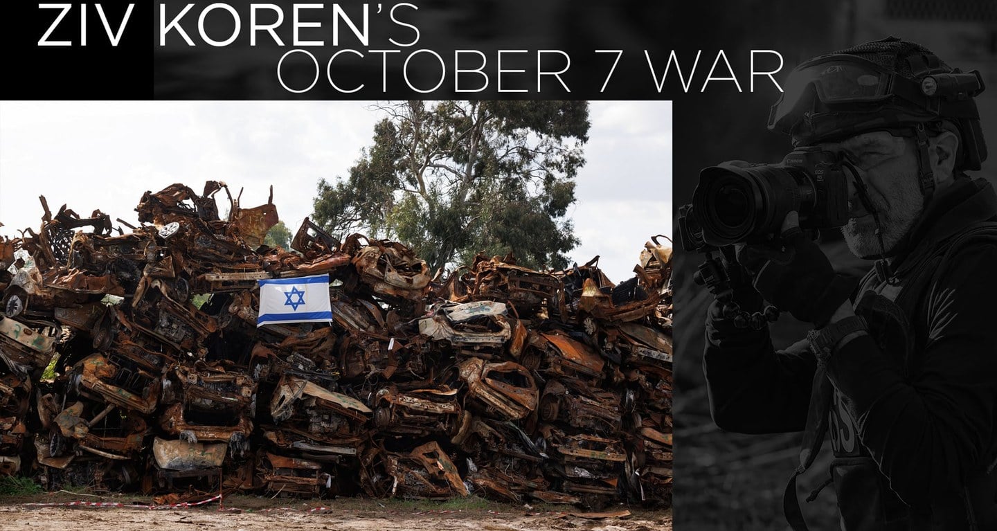 A group of damaged vehicles with an Israeli flag nearby, as a photographer documents the scene on the right side.