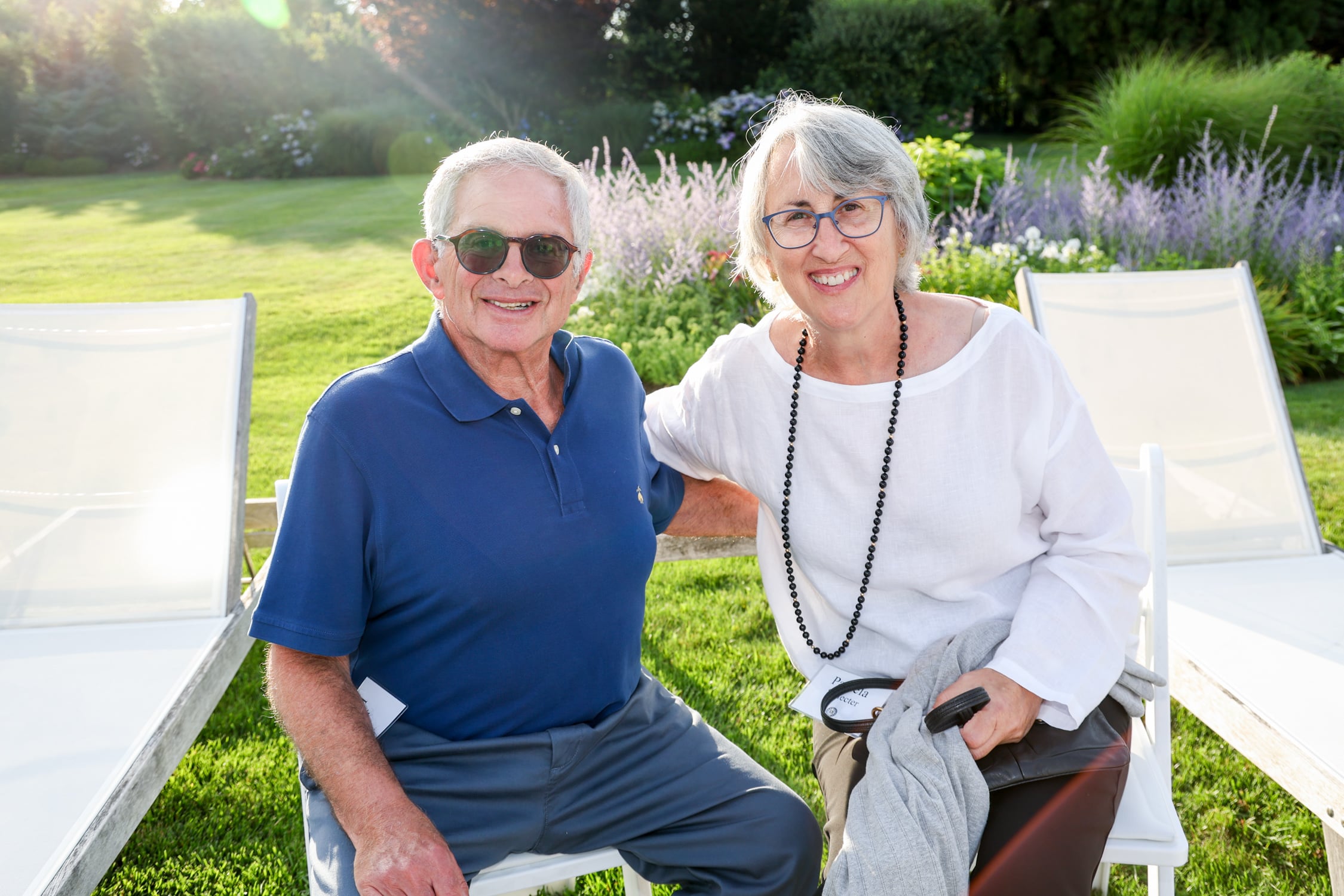 Two smiling older adults sit together on white chairs in a sunlit garden with vibrant greenery and flowers, enjoying a peaceful moment in the outdoor space of Temple Emanu-El.