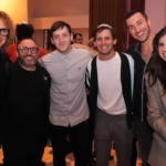 Six community members stand closely together, smiling at a Temple Emanu-El gathering, with other attendees in the background.
