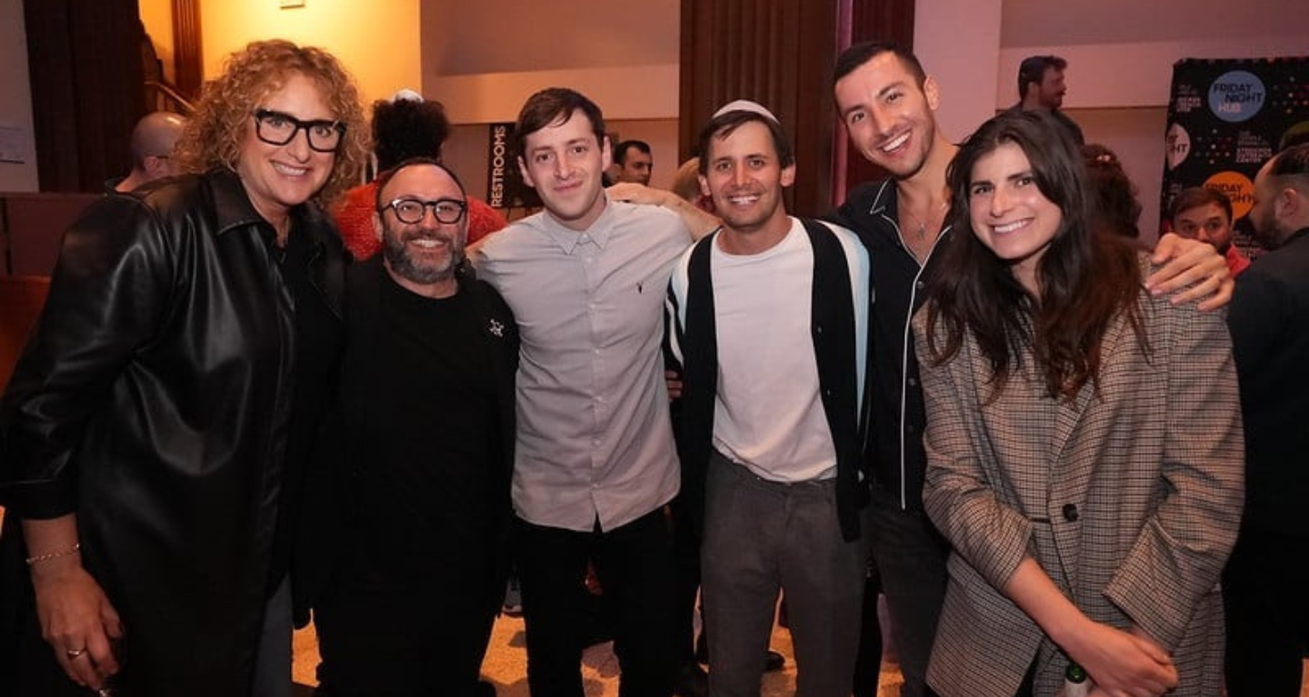 Six community members stand closely together, smiling at a Temple Emanu-El gathering, with other attendees in the background.