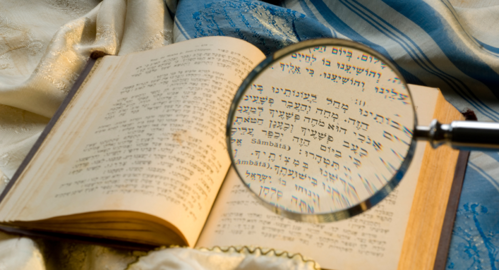 A magnifying glass rests on the open pages of a Hebrew text, with a blue and white tallit draped in the background.