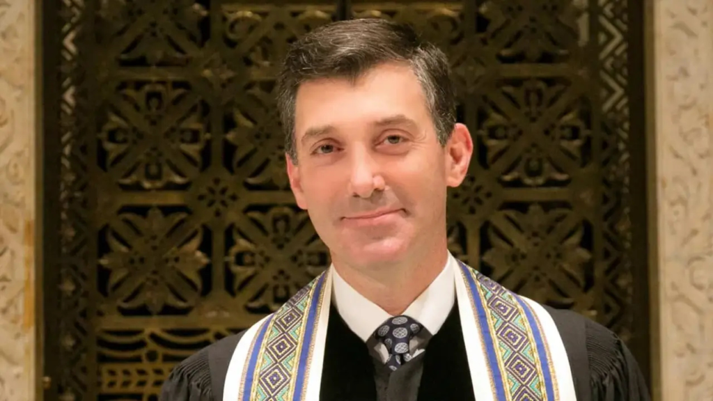 Man dressed in formal attire stands before an elegant, decorative background at Temple Emanu-El, facing the camera.