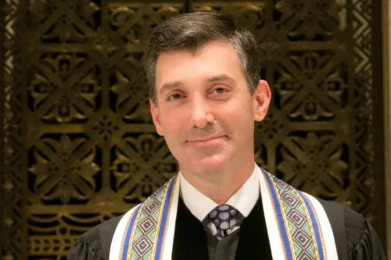 Man dressed in formal attire stands before an elegant, decorative background at Temple Emanu-El, facing the camera.