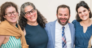 Four congregants smiling and posing together at Temple Emanu-El, dressed in semi-formal, colorful clothing against a light background.