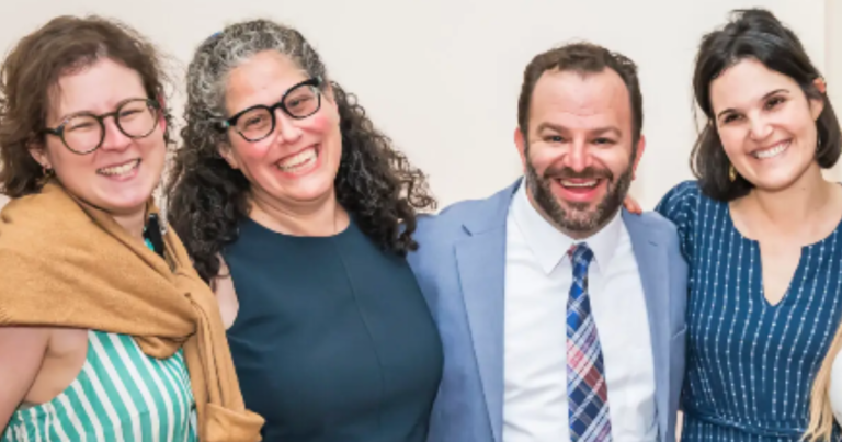 Four congregants smiling and posing together at Temple Emanu-El, dressed in semi-formal, colorful clothing against a light background.