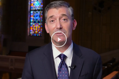Man in suit speaking in a synagogue with stained glass window in background, play button overlay on image.