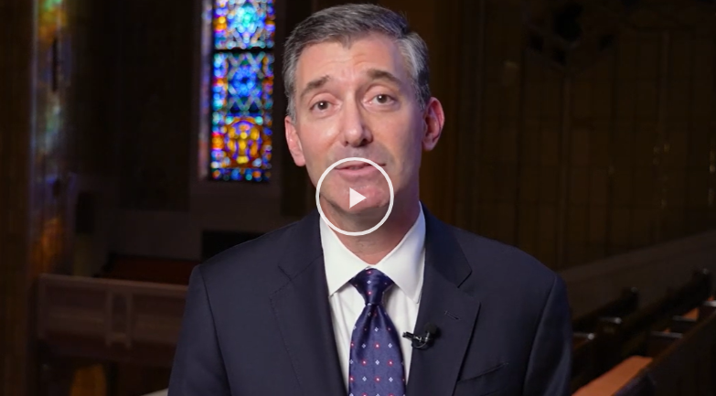Man in suit speaking in a synagogue with stained glass window in background, play button overlay on image.