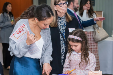 A woman holds bingo sheets and gestures as a girl writes, with others observing during a community gathering at Temple Emanu-El.
