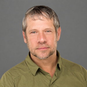 Man with short gray hair and beard, wearing a green shirt, standing in front of a plain gray background at Temple Emanu-El.