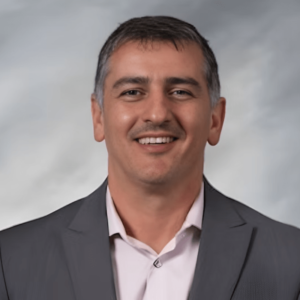 A man in a gray suit and light shirt warmly smiling at the camera, posed against a soft gray background.