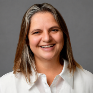 Smiling woman with shoulder-length brown hair, wearing a white blouse, stands against a neutral gray background.