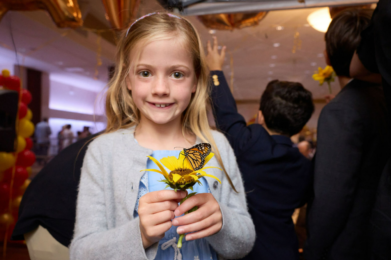 Smiling girl holds a yellow flower with a butterfly on it at a community gathering inside Temple Emanu-El, with other attendees visible in the background.