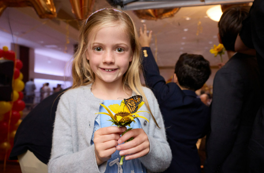 Smiling girl holds a yellow flower with a butterfly on it at a community gathering inside Temple Emanu-El, with other attendees visible in the background.