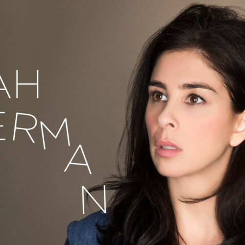 Woman with long dark hair touching her ear, looking upward. Text on the left reads: Sarah Silverman. Simple, neutral background.