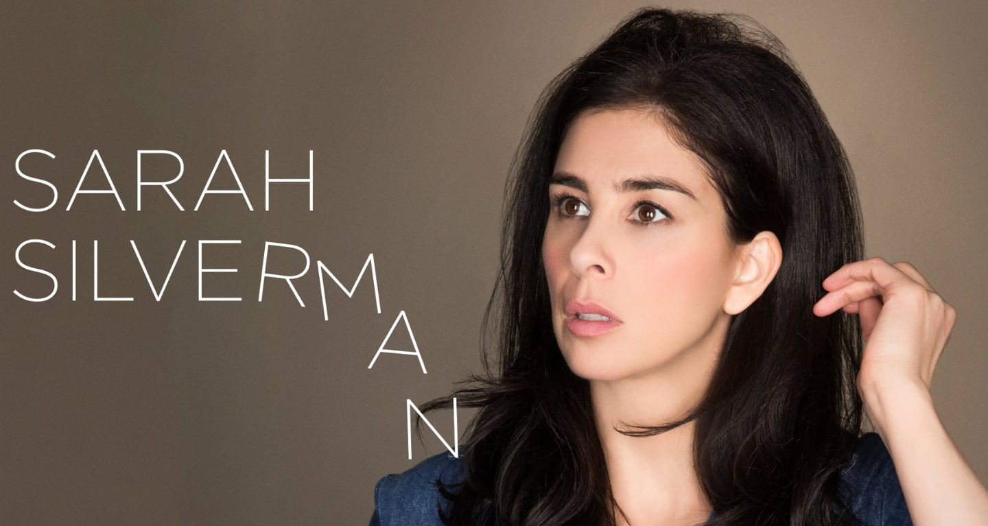 Woman with long dark hair touching her ear, looking upward. Text on the left reads: Sarah Silverman. Simple, neutral background.