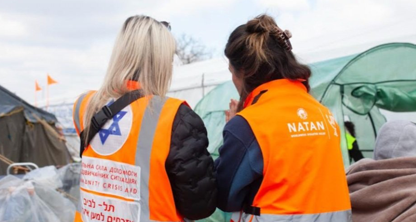 Two aid workers wearing orange safety vests stand near temporary tents at a relief site, seen from behind.