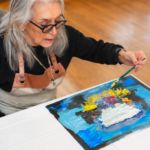 An older woman wearing glasses creates a vibrant abstract painting on a canvas at a table during an art activity at Temple Emanu-El.