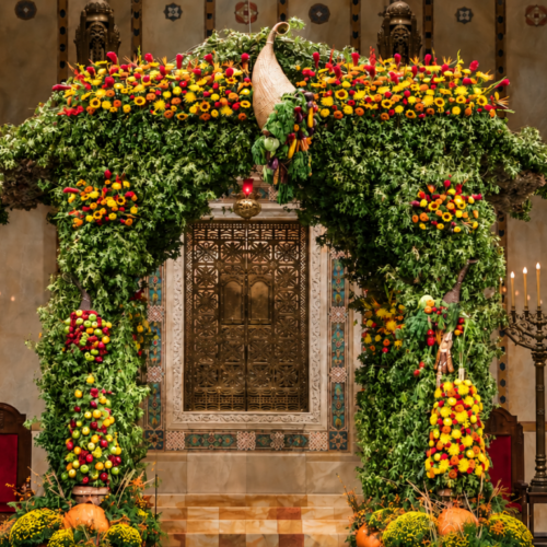 A spacious, beautifully decorated sukkah adorned with green leaves, yellow and orange flowers, and hanging fruit inside Temple Emanu-El in New York City.
