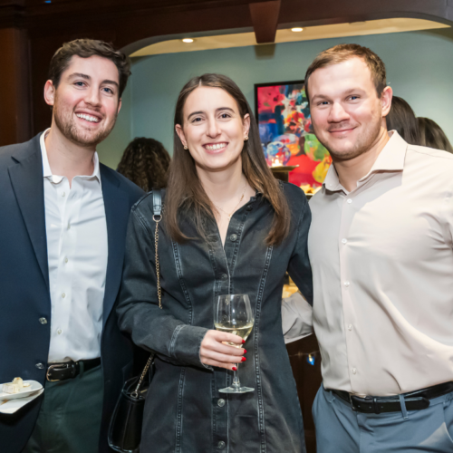 Three people smiling at a Temple Emanu-El gathering, one holding a plate, another with a glass, with others mingling in the background.
