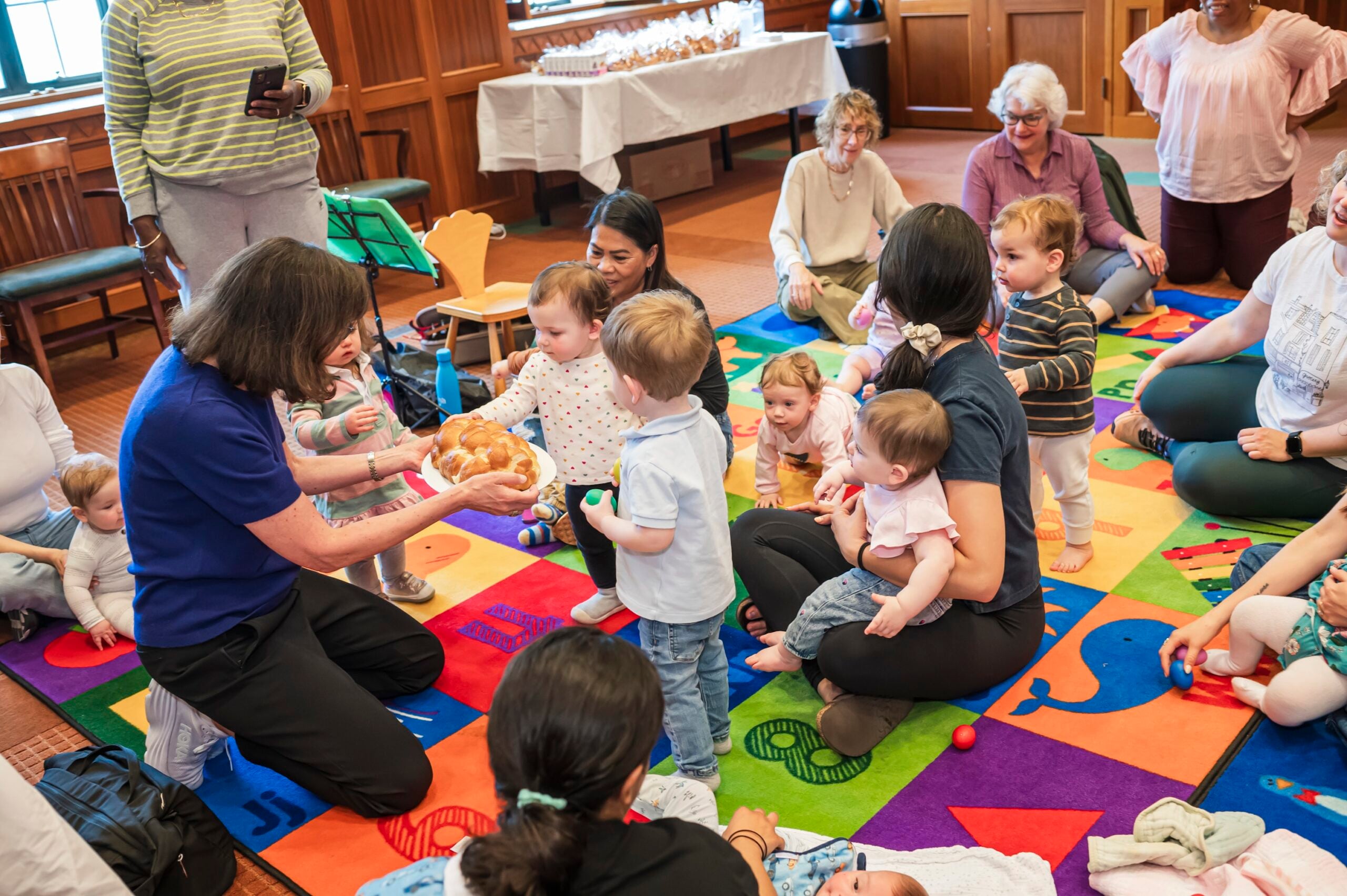 A teacher shares a challah loaf with young children gathered on a colorful play mat at Temple Emanu-El.