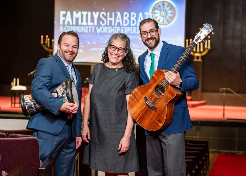 Three Temple Emanu-El clergy members smiling in front of a Family Shabbat screen, two holding musical instruments.