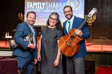 Three Temple Emanu-El clergy members smiling in front of a Family Shabbat screen, two holding musical instruments.