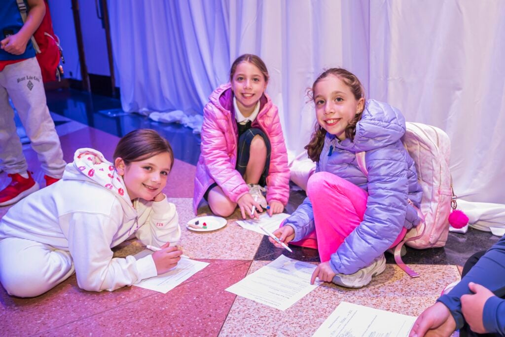 Three young students from the Temple Emanu-El community smiling while sitting on the floor drawing and working on papers.