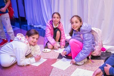Three young students from the Temple Emanu-El community smiling while sitting on the floor drawing and working on papers.