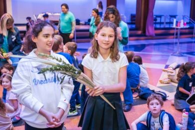 Two girls stand together, one holding greenery, as children and adults gather in a vibrant room at Temple Emanu-El.