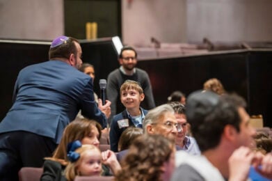 A boy wearing a suit and kippah smiles while speaking into a microphone held by a man at Temple Emanu-El.