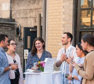 Five people stand around a tall table outdoors on a rooftop at Temple Emanu-El, enjoying conversation and refreshments during a community gathering.