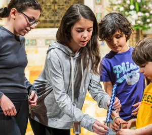 Four children participate in lighting a Havdalah candle together indoors at Temple Emanu-El, with flowers and chairs visible in the background.