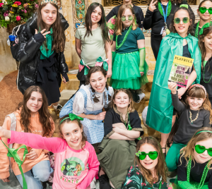 Group of girls in green outfits smiling and posing together indoors at a Temple Emanu-El event, some wearing heart-shaped glasses and holding festive props.