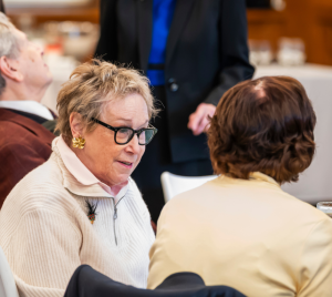 Elderly women engaged in conversation at a table during a formal gathering at Temple Emanu-El, with refreshments and attendees nearby.