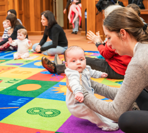 Toddler with a white bow enjoying challah while sitting on a colorful alphabet mat inside Temple Emanu-El.