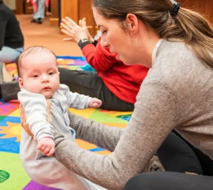 A woman holds a baby upright on a colorful play mat during a group gathering at Temple Emanu-El, with others in the background.