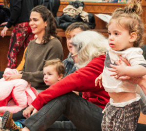 Two adults engage with a baby on a blanket while another adult gently watches over an infant nearby in a welcoming setting.