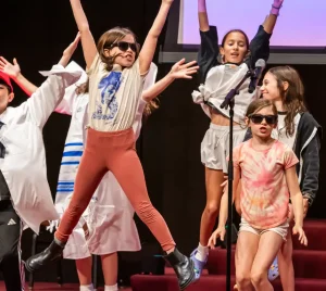 Children on stage at Temple Emanu-El performing energetically; some wear sunglasses and jump with arms raised, others stand smiling.