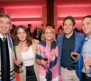 Five people smile at a formal Temple Emanu-El event, holding beverages, with pink lighting in the background.