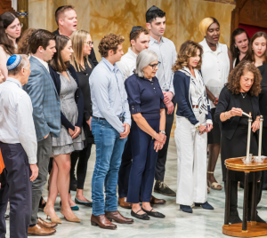 A group of congregants gathers in Temple Emanu-El as a woman lights two Shabbat candles on a small table.