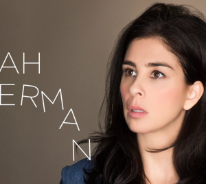 Woman with long dark hair touching her ear, looking upward. Text on the left reads: Sarah Silverman. Simple, neutral background.