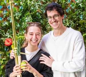 Two congregants smiling in front of lush greenery, one holding a lulav and etrog in celebration of Sukkot at Temple Emanu-El.