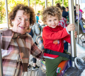 An older woman and a young child share joyful smiles while riding a carousel together on a sunny day, celebrating special moments of community and connection.
