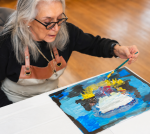 An older woman wearing glasses creates a vibrant abstract painting on a canvas at a table during an art activity at Temple Emanu-El.